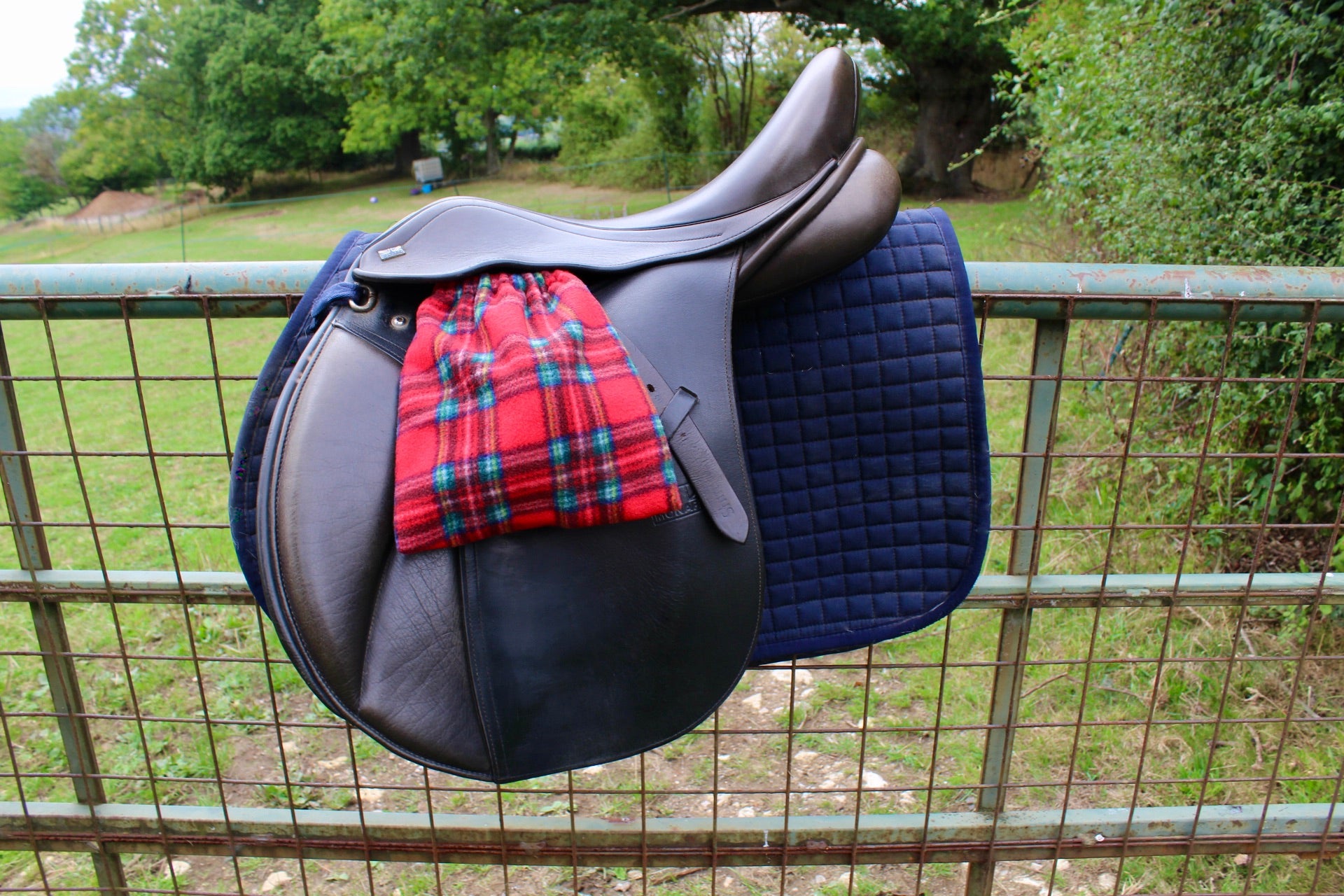 red tarten stirrup covers on a brown monarch saddle which has a navy le Mieux saddle pad under, on a field fence