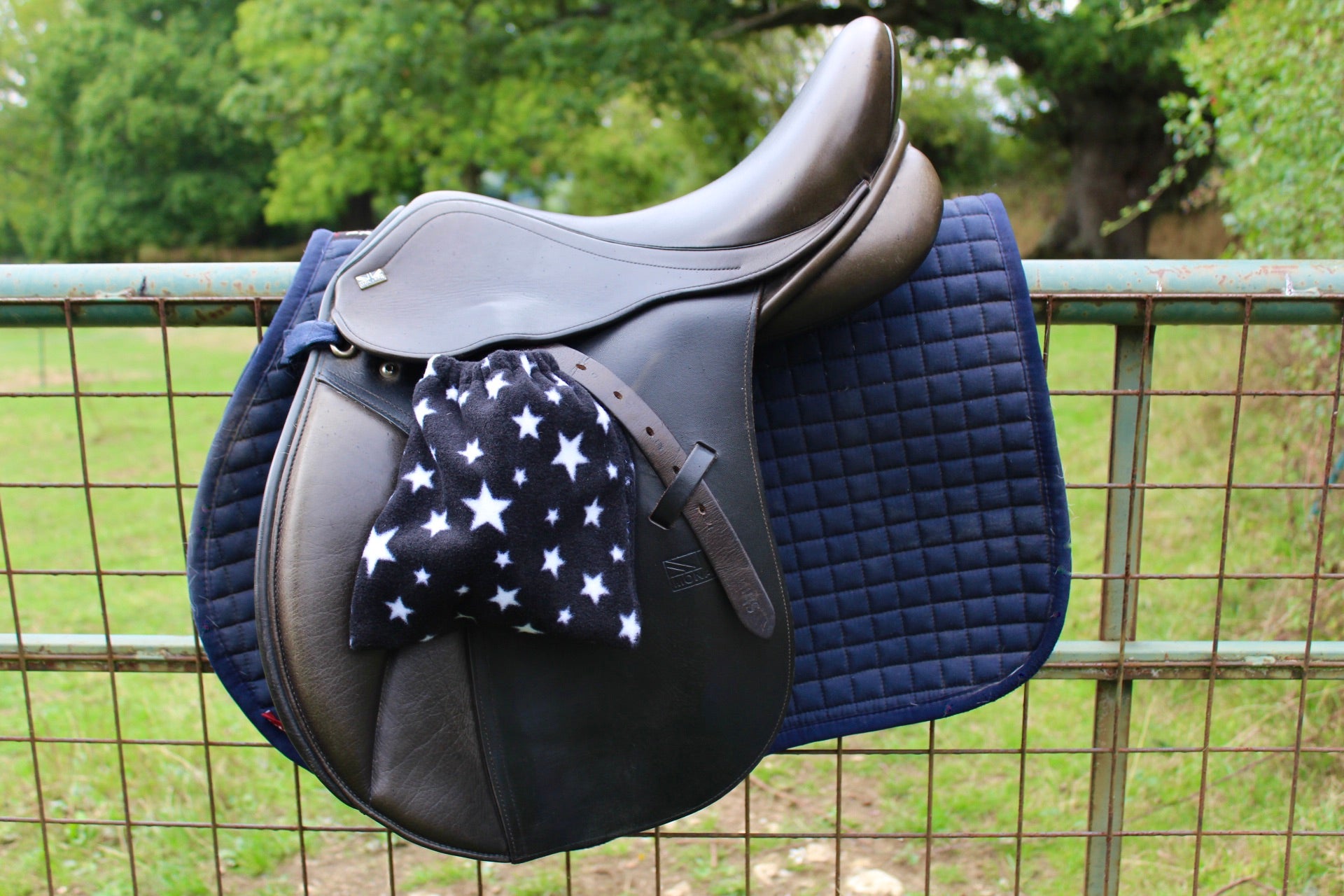 black stirrup covers with white stars on them, on a brown monarch saddle which has a navy le Mieux saddle pad under, on a field fence