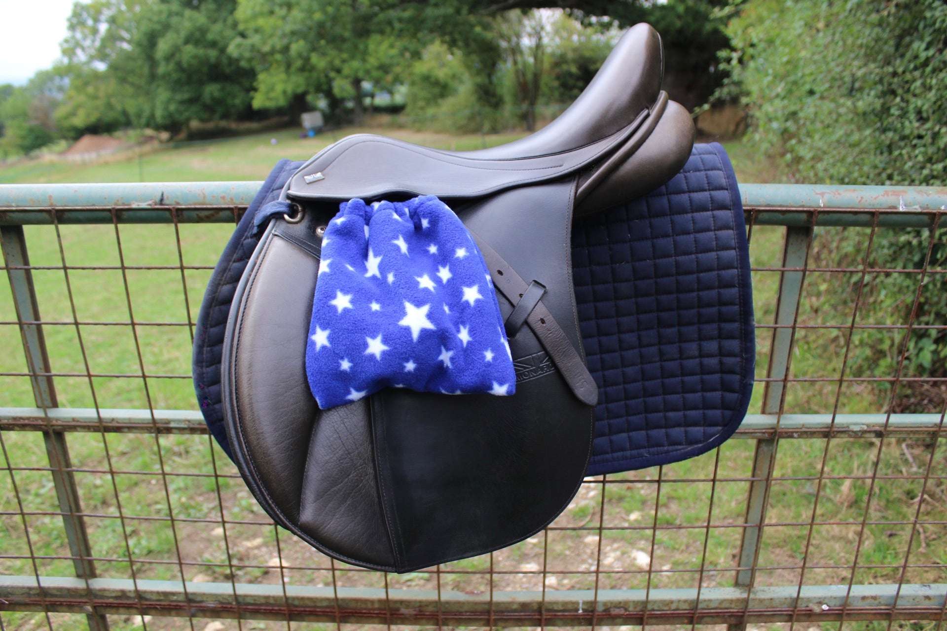 blue stirrup covers with white stars on them, on a brown monarch saddle which has a navy le Mieux saddle pad under, on a field fence