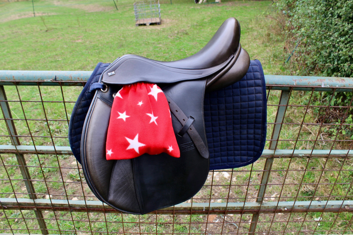 red stirrup covers with white stars on them, on a brown monarch saddle which has a navy le Mieux saddle pad under, on a field fence