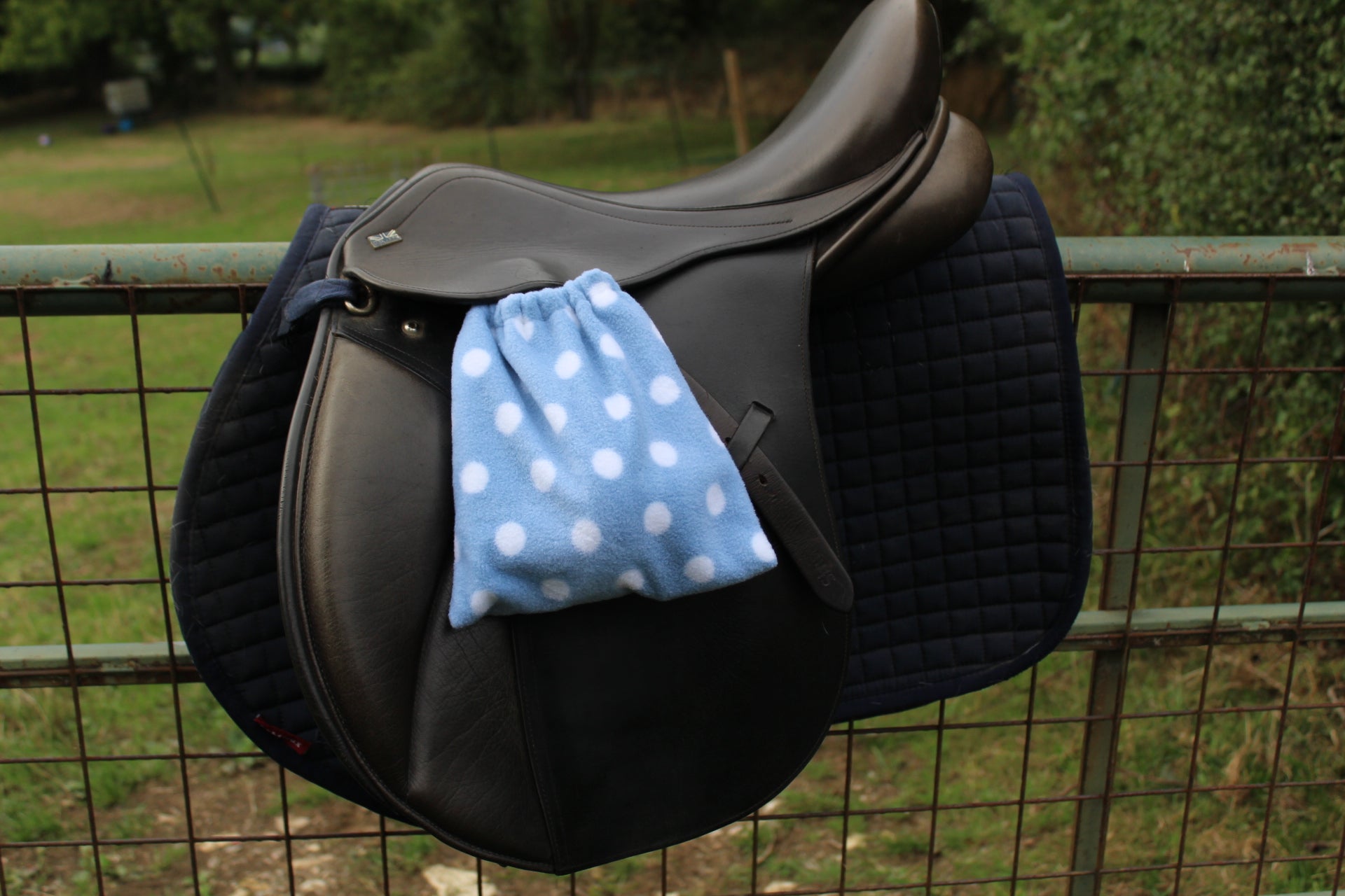 light blue stirrup covers with white spots on them, on a brown monarch saddle which has a navy le Mieux saddle pad under, on a field fence